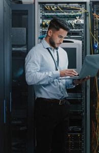 IT professional using a laptop while working beside server racks in a data centre